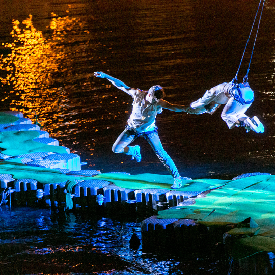 Two people on a platform above water, one person is suspended in the air. Illuminated by blue and green lighting.